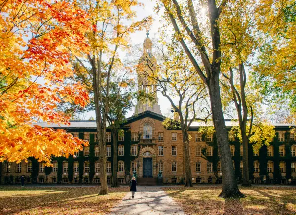 A student walking past a historic college building framed by autumn trees.
