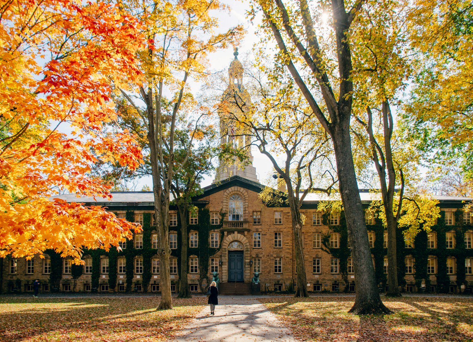 A student walking past a historic college building framed by autumn trees.