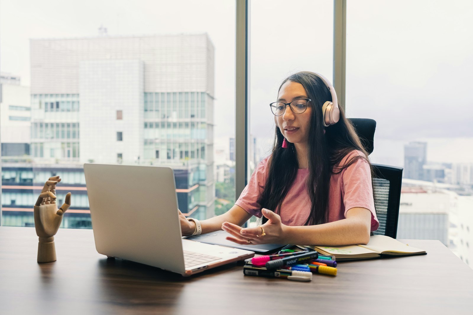 A student wearing headphones speaking to her laptop at a desk by a high-rise window.