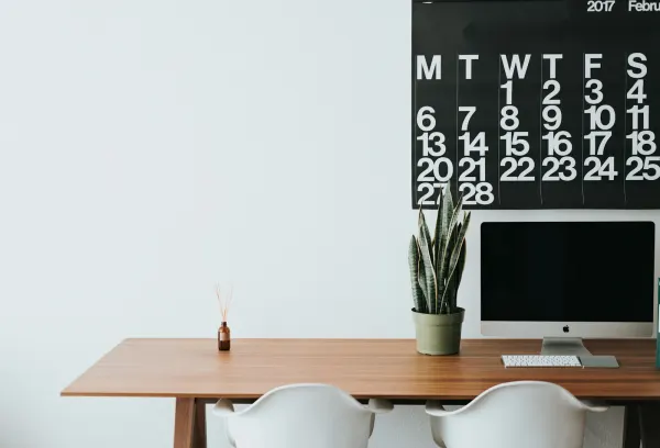 A large wall calendar above a tidy desk with a computer and a plant.