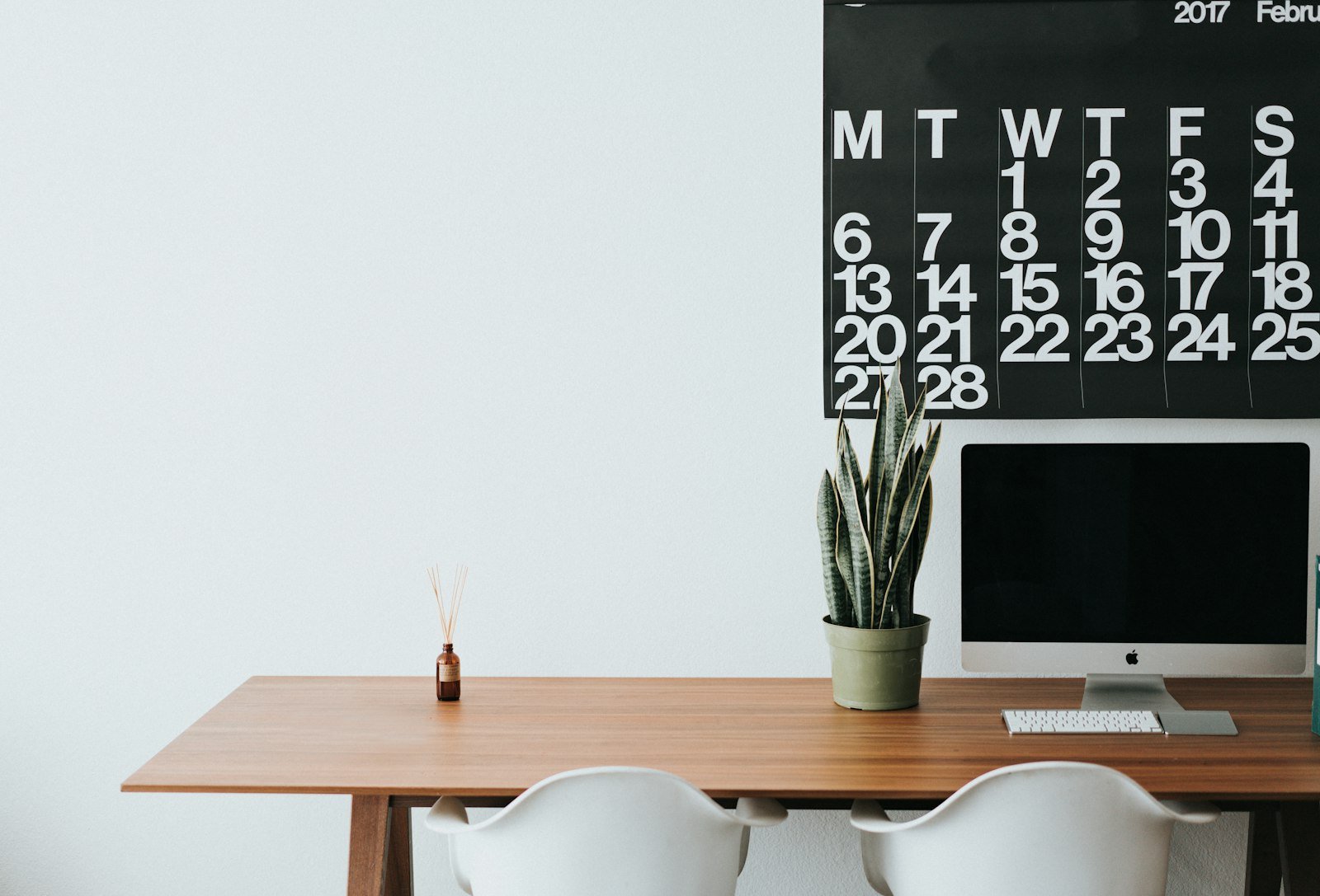 A large wall calendar above a tidy desk with a computer and a plant.